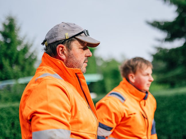 Ein älterer und ein jüngerer Arbeiter in jeweils orangefarbener Arbeitskleidung schauen nach rechts in die Ferne.