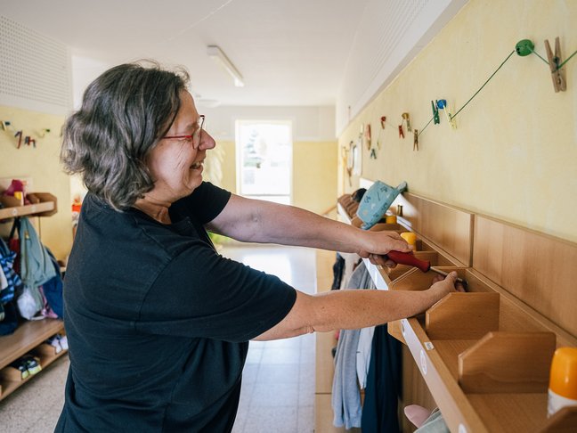 Eine in schwarz gekleidete ältere Frau mit Brille und grauen Haaren schraubt mit einem Schraubenzieher an einem Holzregal im Flur eines Kindergartens.