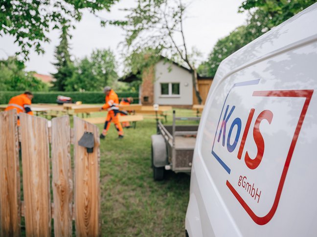 Zwei Mitarbeiter der KOIS gGmbH stellen in einem Garten Holzbänke auf. Rechts im Vordergrund steht ein weißer Transporter mit Anhänger, welcher die blaurote Aufschrift KOIS trägt.