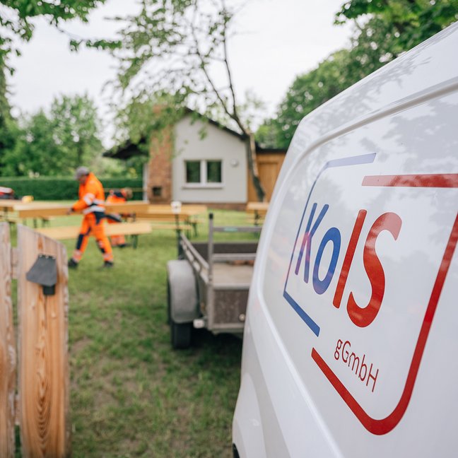Zwei Mitarbeiter der KOIS gGmbH stellen in einem Garten Holzbänke auf. Rechts im Vordergrund steht ein weißer Transporter mit Anhänger, welcher die blaurote Aufschrift KOIS trägt.