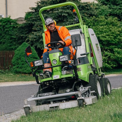 Ein Mann in orangefarbener Arbeitskleidung sitzt auf einem grünen Mähfahrzeug und mäht eine Wiese. 