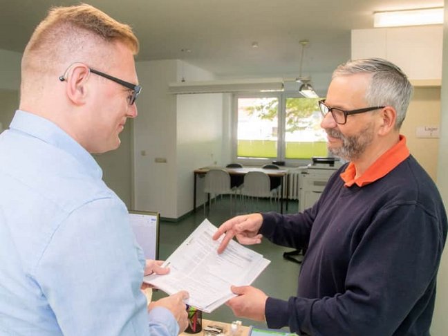 Ein rechts stehender Mann mit einer Brille und in einem blaugrauen Pullover zeigt einem links stehenden Mann im blauen Hemd und mit einer Brille, ein Dokument. Die beiden befinden sich in einem Büroraum. 