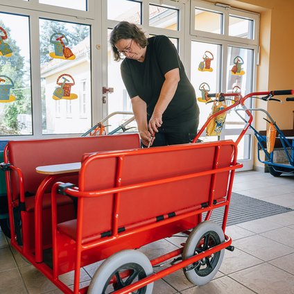 Eine in schwarz gekleidete ältere Frau mit Brille schraubt mit Werkzeug an einem roten Bollerwagen im Flur eines Kindergartens.
