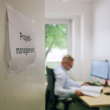 Die Tür zu einem Büroraum steht offen. Links an der Tür befindet sich ein Schild mit der Aufschrift: Projektmanagement. Im Raum sieht man einen Mann mit Hemd und Brille am Computer sitzen mit Dokumenten in der Hand arbeiten. 