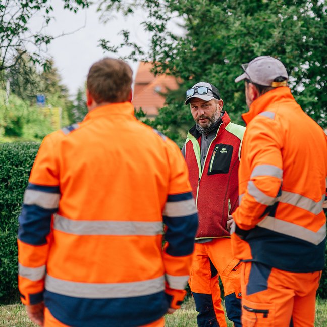 Drei Männer in organgefarbener Arbeitskleidung sprechen miteinander