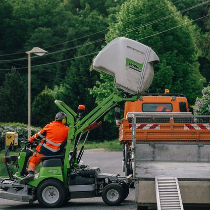 Ein Mann in orangefarbener Arbeitskleidung sitzt auf einem grünen Mähfahrzeug und kippt das gemähte Heu auf einen Anhänger hinter der Ladefläche eines orangefarbenen Transportfahrzeugs.