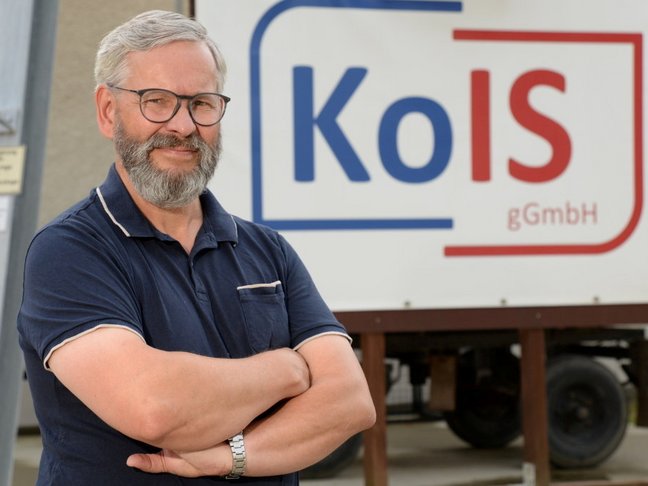 Ein Mann mit grauen Haaren, Bart und Brille, in einem blauen Poloshirt steht mit verschränkten Armen neben einem Schild mit der Aufschrift Kois, von der Kodersdorfer Inklusions gGmbH. 