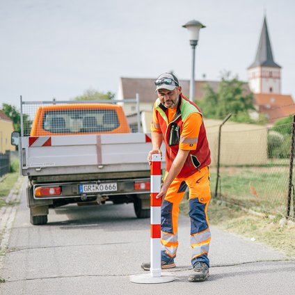 Ein Arbeiter in orangefarbener Arbeitskleidung stellt einen Poller in der Mitte einen kleinen Weges auf. Im Hintergrund befindet sich sein organgenes Arbeitsfahrzeug mit grauer Ladefläche.