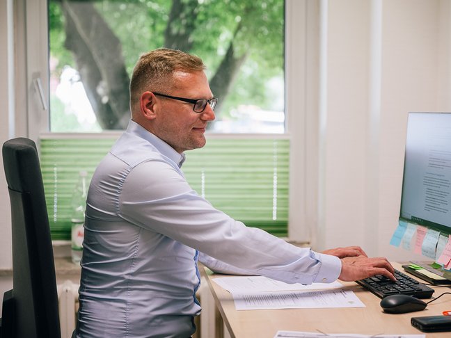 Ein junger Mann mit Brille und einem blauen Hemd sitzt in einem Büroraum vor einem Computer und tippt mit seinen Fingern etwas auf der Tastatur.