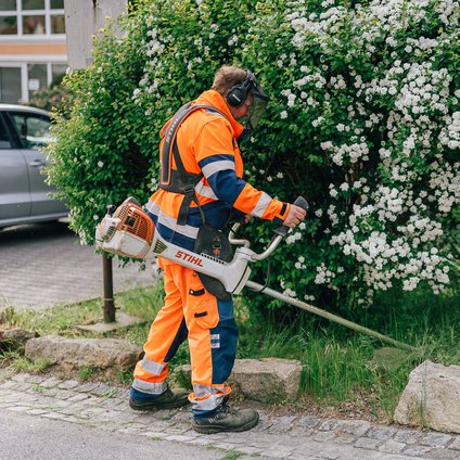 Ein Arbeiter in orangefarbener Arbeitskleidung mäht mit einer Motorsense eine Wiese.