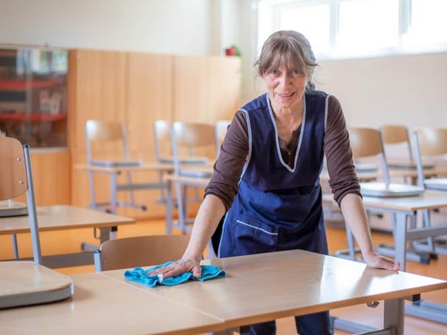 Eine alte Frau putzt einen vorn im Klassenzimmer stehenden Tisch mit einem Putzlappen und schaut dabei freundlich in die Kamera.