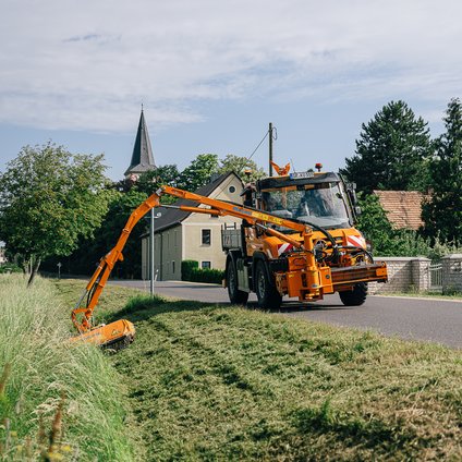 Ein orangefarbenes Mähfahrzeug mäht das Gras an einem Straßenrand mit einem langen Greifgerät.