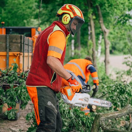 Ein Mann in roter Weste, Schutzhelm und Arbeitshose steht mit einer Kettensäge in den Händen haltend im Vordergrund des Bildes umgeben von abgesägten Ästen. Im Hintergrund sammelt ein Mann mit orangefarbener Arbeitskleidung die Äste auf. Links im Hintergrund ist ein orangefarbenes Arbeitsfahrzeug mit Ladefläche zu sehen. 